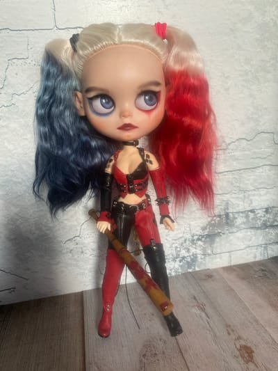 This media file shows a doll resembling Harley Quinn from DC Comics. The doll has blonde hair with red streaks and blue streaks, and is wearing a red and black outfit with studded accents. The doll is holding a baseball bat. The background appears to be a surface with a brick pattern and a wooden texture. The lighting suggests an indoor setting, likely during the daytime. No text is visible. The doll is positioned lying on its back, with its head slightly tilted. The overall presentation is that of a collectible doll display.
