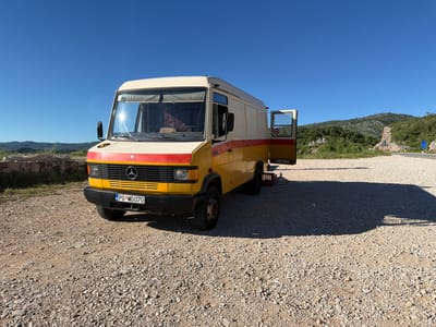 A yellow and white van with red stripes is parked on a gravel road in a rural area with hills and a clear blue sky. The van has its side door open, revealing a metal grate. A speed limit sign of 80 is visible on the open door. The license plate of the van reads "PG*MG 070". The scene appears to be in Ponikvica, Montenegro, indicated by the license plate format.