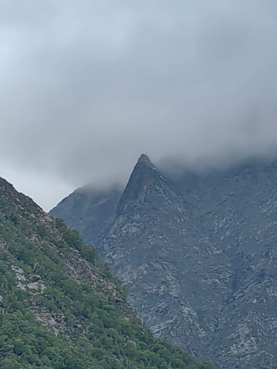 The image shows a mountainous landscape under a cloudy sky. In the foreground, a steep, rocky slope covered in green vegetation, likely trees and bushes, recedes diagonally from the bottom left towards the upper right. Behind this slope, several jagged mountain peaks rise. One prominent peak in the center, with a sharp, pointed summit, is partially obscured by clouds. The mountains are rugged and appear to be composed of dark gray rock. The overall atmosphere is somber and dramatic, suggesting a cool, overcast day in a high-altitude region. The scene is in the vicinity of Gondhla, India, a location known for its dramatic mountain scenery. There are no people or man-made structures visible in the image, indicating a natural, remote environment.