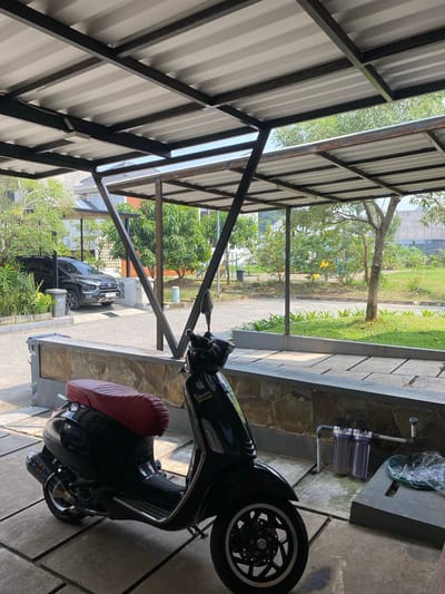 The image shows a black Vespa scooter with a red seat parked under a corrugated metal and metal-framed awning in Cibinong, Indonesia. The awning has a light gray, ribbed roof supported by dark metal beams. The ground beneath the awning is paved with large concrete tiles. To the left of the scooter, a black SUV is visible parked in the background, near a row of leafy green trees and a modern building with white and orange accents. A gray trash bin is situated next to the SUV. Beyond the awning, a neatly maintained lawn with scattered trees and shrubs leads to a paved area and a building in the distance. In the foreground, a low stone wall borders a raised garden bed. Next to the scooter, a water filtration system with two clear canisters is mounted on the wall. The lighting suggests it is daytime with bright sunlight.