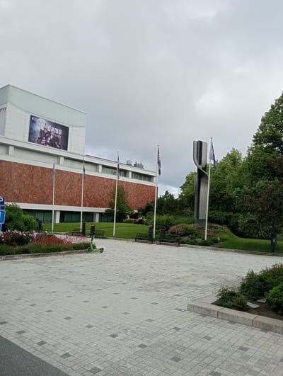 This media file depicts an outdoor scene in Kuopio, Finland, likely during the daytime with overcast weather. The foreground is a paved plaza with a repeating pattern of light gray and darker gray square pavers. A bright green electric scooter is parked on the plaza.

Behind the plaza, there's a landscaped area with green grass, bushes, and flowerbeds filled with red and pink flowers. Several park benches are visible along the edge of the grass. A modern, abstract metal sculpture stands prominently in the middle ground, flanked by two flagpoles with dark blue flags. Behind the sculpture and landscaping, a large, modern building with a brick facade and a lighter colored upper section is visible. A large banner advertising "The Adams Family" is displayed on the upper part of the building, suggesting a theatrical production.

The overall atmosphere is calm and perhaps a bit subdued due to the cloudy sky. The presence of the building with the banner, sculpture, and paved public space suggests this is a cultural or civic area within the city. No people are clearly visible in the frame, focusing the viewer's attention on the architecture, landscaping, and the sculpture. The lighting is diffused, consistent with an overcast day.