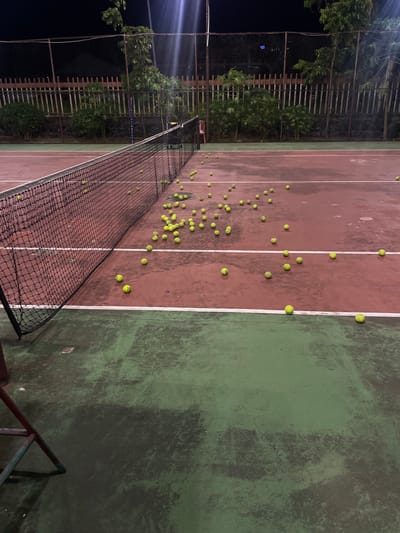 The image depicts an outdoor tennis court at night, likely located in Manado, Indonesia, based on the provided context. The court surface is a dark red with green sections. Numerous yellow tennis balls are scattered across the court, many near the net and extending into the playing area. The net is black and stretched across the width of the court. In the background, a dark fence and lush green trees are visible, with artificial lighting casting shadows and creating a somewhat moody atmosphere. There are no people visible in the scene. The court appears somewhat worn, with some patches and scuff marks visible on the surface.