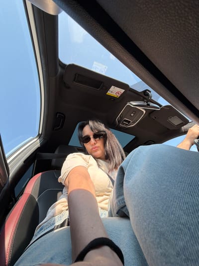 In the city of Fyodorovo, Russia, a woman is captured from a low angle, inside a car. She is sitting in the driver's seat, her left arm resting on her lap. The sun shines brightly through the open sunroof, casting a warm glow on her. She wears a light-colored t-shirt and blue jeans, with sunglasses completing her look. The car's interior is visible, including the dashboard, a rearview mirror, and the sun visors. The seats appear to be made of black and red leather. The overall scene suggests a pleasant day, with the woman enjoying a drive.