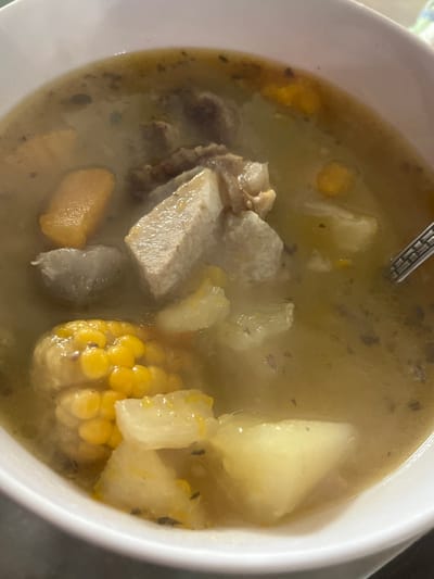 A close-up shot shows a bowl of hearty soup, likely a sancocho, a traditional Venezuelan stew. The soup is filled with various ingredients including chunks of meat, pieces of potato or yuca, and a segment of corn on the cob. The broth is a yellowish-brown color, speckled with herbs and spices. A silver spoon is partially visible on the right side of the bowl, suggesting someone is about to eat. The background is blurred, but appears to be a light-colored surface, possibly a table. The image is framed to focus entirely on the soup, highlighting its rich texture and appetizing appearance. The image does not provide any information about its location in San Antonio, Venezuela.