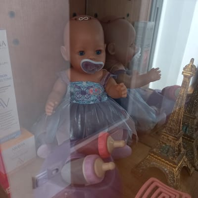 The image shows a baby doll wearing a blue and silver dress with butterfly motifs and a pacifier. The doll is positioned inside a display case, reflected in the glass. To the left of the doll, there are cosmetic product boxes. To the right, a miniature golden Eiffel Tower replica is visible. In the foreground, two baby bottles are present. The setting appears to be a shop or a home display in Cepeleuți, Moldova. The lighting is dim, and the image is slightly blurry due to reflections.