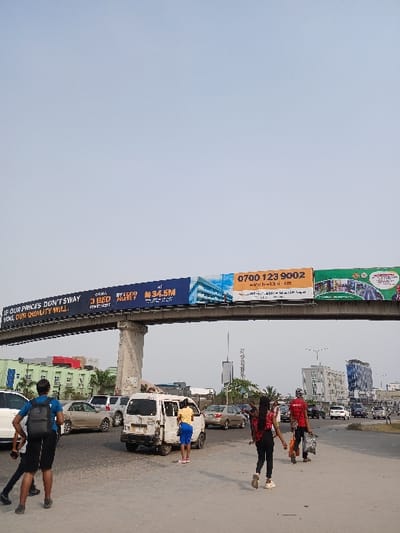 The image displays an urban street scene in Nigeria under a clear, bright sky. A prominent feature is a long, elevated advertising billboard structure, supported by concrete pillars, stretching across the frame above the road.
Several people are present on or near the road. In the foreground, a person with a black backpack walks towards the left. Further into the scene, a person in a yellow shirt and blue shorts stands next to a white van, appearing to interact with it. To their right, two individuals are walking, one wearing a red checkered shirt and the other a plain red shirt.
The road is busy with several vehicles, including various cars and the white van, which seems to be stationary. In the background, multiple buildings line the street, contributing to the urban setting.
The large billboard structure features several advertisements with visible text. One section reads, "IF OUR PRINCES DON'T SWAY YOUL, OUR QUALITY WILL." Other parts of the billboard display "3 BED" and "AT ₦34.5M," indicative of real estate advertising. Contact information includes a phone number, "0700 123 9002," and a web address, "www.h-wick.com" (as per OCR "h-wick em"). The overall scene depicts a typical daytime street activity in a bustling city.
