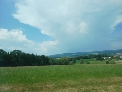 This is a landscape photograph taken in Oberviechtach, Germany. The scene depicts a vast, grassy field in the foreground, with a dense line of dark green trees forming the midground. Beyond the trees, rolling hills fade into the distance, dotted with patches of greenery and scattered buildings, suggesting a rural or semi-rural setting. In the far distance, a town or village is visible on the right side of the frame, with houses and rooftops. The sky above is a mix of bright blue and dramatic white clouds, with some darker clouds towards the right side, indicating potentially changing weather. The overall impression is one of a serene, natural environment under a dynamic sky. There is no visible text or activity taking place, and the time of day appears to be daytime, likely afternoon, given the lighting.