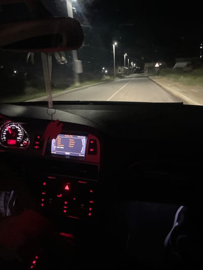 The image shows the interior of a car at night, driving on a road with streetlights and some buildings visible. The dashboard is illuminated, showing the speedometer, a car radio display, and various control buttons. A red air freshener hangs from the rearview mirror. The road ahead is paved and marked with white lines. The general environment appears to be a suburban or rural area at night, with low-level lighting. The location is specified as Valkosel, Bulgaria. There are no people visible in the frame, only the interior of the car and the external environment as seen through the windshield.