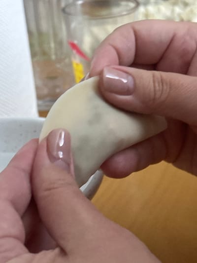A close-up shot captures two hands actively engaged in preparing food, likely shaping a dumpling. The hands are holding a single, crescent-shaped dumpling with a pale, possibly wheat-based wrapper that appears slightly translucent, revealing a darker filling inside. The fingers are delicately pinching the edges of the dough to seal or pleat the dumpling. Both hands have manicured fingernails, painted with a glossy, light beige or nude-colored polish.

The background is softly blurred, indicating a shallow depth of field and drawing focus to the hands and the food item. Hints of a kitchen or dining environment are visible in the background, including vague shapes of clear containers or glassware and a white surface, possibly a paper towel or tablecloth. The overall scene suggests a domestic setting during food preparation. No specific time of day or weather conditions can be inferred, as the activity is indoors. There is no visible text in the image. The scene is located in Egkomi Lefkosias, Cyprus.