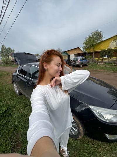 A young woman with red hair, wearing a white shirt, stands outside next to a black car. She is smiling and looking off to the side with her hand near her face. The car is on a grassy area, likely a driveway or roadside. In the background are several buildings, including a yellow one. Another car, possibly a police car, is visible in the distance. The sky is overcast. The location is Chastyye, Russia.