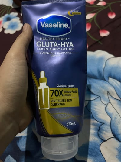 A hand holds a blue and yellow tube of Vaseline Gluta-Hya Serum Burst Lotion, 330ml. The lotion tube features text indicating "HEALTHY BRIGHT™", "GLUTA-HYA", "SERUM BURST LOTION", and "OVERNIGHT RADIANCE REPAIR". There is also an illustration of a dropper bottle and text stating "GlutaGlow • Hyaluron", "70X Melatonic-Peptide Complex*", and "REVITALISES SKIN OVERNIGHT". The background is a blurry floral patterned fabric with blue and white flowers. The image is undated and location is unknown, however, the user stated that this is in Banyusri, Indonesia.