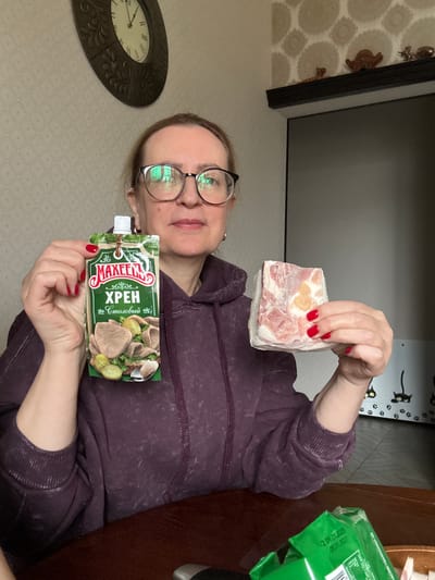 A woman is depicted indoors, likely within a home setting in Krapivka, Russia. She is wearing glasses and a dark purple long-sleeved top. The image, which appears to be a selfie, shows her presenting two food items.
In her right hand, she holds up a rectangular block of what seems to be frozen meat or lard, characterized by layers of pinkish meat and white fat, with a small yellowish portion visible. Her fingernails are painted bright red. In her left hand, she displays a green flexible pouch of "МАХЕЕВ ХРЕН Столовый" (Makheev Table Horseradish). The packaging features an illustration of sliced meat, possibly tongue, served with pickled cucumbers and dill.
The background consists of a light-colored wall covered in wallpaper with a subtle, repeating geometric pattern. To her left, a partially visible, ornate dark-framed wall clock with Roman numerals indicates a time near the top of the hour. Above her right shoulder, a dark-framed item, possibly a screen or mirror, is visible, with small decorative figurines positioned on a shelf above it. To her right, a dark wooden table holds a green package with Russian text, including dates "2.09.01.2008" and "08.01.2027", along with words like "ПОЛОЦКИИ" (Polotskiy) and "ЭКСТР" (Extra). A decorative element featuring black cat silhouettes and paw prints adorns the wall in the upper right. The woman maintains a neutral expression, and the scene suggests a domestic activity such as preparing or presenting food.