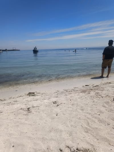 The image depicts a serene beach scene under a clear blue sky with wispy clouds. In the foreground, a person is standing on a sandy beach, facing away from the camera, looking out at the calm, clear water. The person is wearing a black t-shirt and tan shorts. The water is shallow near the shore, revealing the sandy bottom. Further out in the water, a small boat is visible, and in the distance, a few individuals are engaged in water activities, possibly paddleboarding or jet skiing. A pier structure is also discernible on the left side of the image. The overall atmosphere is tranquil and suggests a pleasant day at the beach. The location is identified as Oakville, Canada.