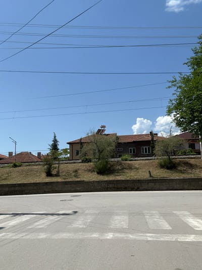 The image depicts a street in Kresna, Bulgaria. Buildings are visible along one side of the street, with power lines overhead. A crosswalk is painted on the asphalt road. The architecture of the buildings suggests a residential area. Vegetation is present along the edge of the road, indicating some landscaping or natural growth. The sky is clear and blue.