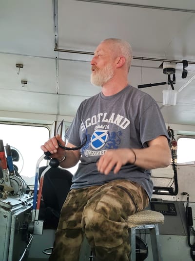 A man with a gray beard and a t-shirt that says "Scotland" is at the helm of a vessel. He is wearing camouflage pants and sitting on a cushioned stool. The interior of the vessel is utilitarian, with metal paneling and various controls and communication devices. The image was taken in Anisimovo, Russia.