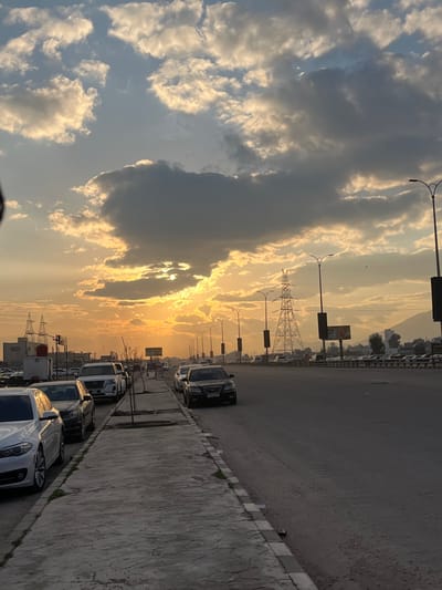The image depicts a wide, multi-lane urban road and an adjacent sidewalk at sunset in Al-Qaboun Municipality, Syria. The sky is characterized by an orange-yellow sunset horizon, obscured by large, dark grey clouds and interspersed with brighter white clouds, with the sun partially visible through a break in the clouds near the center-left.

In the foreground, a concrete sidewalk with visible cracks and some light green vegetation, likely small trees or bushes, runs along the left side of the frame. Several vehicles are parked parallel to the curb on the left. From foreground to background, these include a white BMW 5 Series sedan, a dark-colored sedan (possibly black), and an SUV (possibly white Nissan Patrol), followed by other indistinguishable vehicles. On the right side of the sidewalk, a dirt strip separates it from the main asphalt road.

The asphalt road features multiple lanes, suggesting it is a major thoroughfare. Vehicles are visible on the road, both moving and parked further down. A black Hyundai sedan is parked closer to the foreground on the right side of the road, facing away from the viewer.

Along the road, numerous streetlights with double arms are visible, extending into the distance on both sides. Several tall, metal lattice-work electricity transmission towers (pylons) are prominent features in the middle ground and background, particularly one large tower in the mid-right. In the far background, a range of dark mountains is visible under the fading light. There are also what appear to be billboards or large signs along the right side of the road in the middle distance. No individuals are clearly discernible in the scene.