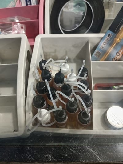 The image shows a collection of small bottles with black and white caps, some with white plastic loops attached. These bottles appear to contain a light amber liquid. They are neatly arranged in a white plastic organizer with multiple compartments. The organizer is placed on a dark, possibly glass, surface with a speckled texture. In the background, other objects are visible, including a pink plastic organizer, a roll of black tape, and what looks like a soldering tool with a gold-colored handle. The scene suggests a workspace or hobby area, possibly for electronics or crafting. There is visible text on some of the items, including on the packaging of the soldering tool and on a small round container in the lower right corner that says "H SOLDER PASTE". The overall lighting is moderate, with no strong shadows, and the setting appears to be indoors. The image is a close-up shot focusing on the bottles and the organizer.