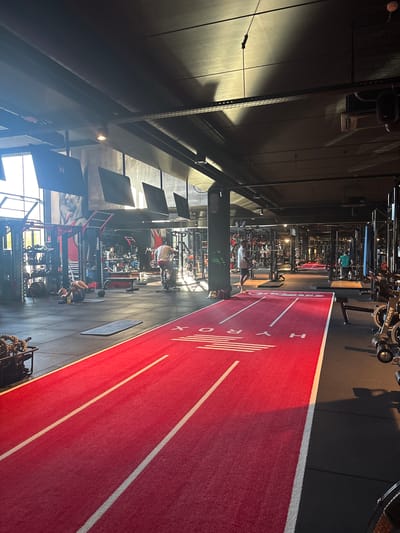 The image depicts the interior of a well-lit, modern gym with a distinctive red running track. The scene is a long shot, showing the length of the gym, with various exercise equipment and machines lining the left side. Several people are visible, engaged in workouts. On the right, the red track, marked with white lines and the word "HYDRO" in large white letters, dominates the foreground and extends into the distance. The gym appears to be busy, with individuals using treadmills, weight machines, and other fitness equipment. Some individuals are in the process of exercising, while others might be resting or transitioning between activities.
Notable details include the presence of specialized gym equipment, possibly for high-intensity interval training given the "HYDRO" branding on the track. The lighting suggests it is daytime, with natural light filtering in from windows on the left, casting shadows and highlighting the textures of the gym floor and equipment. The overall atmosphere seems energetic and focused, typical of a fitness environment.
Visible text includes "HYDRO" on the track. Further in the gym, on the equipment, text like "octagon" and "ESCAPE YOUR LIMITS." is visible on some of the machines. The location context is Destelbergen, Belgium. The image does not convey specific emotions or weather conditions, beyond the active and perhaps determined mood of the gym-goers.
