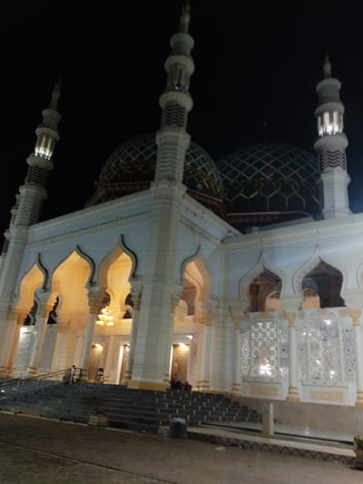The image displays a large mosque at night in Kotapantonlabu, Indonesia. The structure is predominantly white or light-toned, illuminated by internal and external artificial lighting. Two large, dark green domes, featuring a diamond grid pattern and gold-colored decorative elements at their bases, cap the main building. At least three minarets are visible; two flank the main structure (one on the far left, one on the far right), and one is centrally positioned between the domes. These minarets are cylindrical, white, and adorned with red and white zig-zag patterned bands; their upper sections emit light. The mosque's facade presents multiple pointed arches, through which a warm, yellow light is visible from the interior. Pillars support these arched sections. On the lower right side of the building, intricate white latticework panels are integrated into the wall design. A wide, multi-tiered staircase, dark grey in color and equipped with metal handrails, leads up to the mosque's entrance. Two indistinct human figures are discernible on the staircase. The foreground consists of a dark, unpaved surface, with a small potted plant situated near the base of the stairs. The sky above is completely dark, indicative of a nocturnal setting.