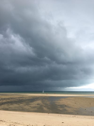 The image depicts a beach in Baan Kai, Thailand. The scene is dominated by a large, dark storm cloud formation covering the left side of the frame. The right side of the frame shows a sandy beach, partially wet from the receding tide. The water appears calm and pale in color. Several small boats or sails are visible on the beach. Two small figures are on the sand.