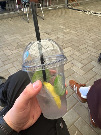 A first-person perspective shot captures a hand holding a clear plastic cup filled with a clear liquid, ice, mint leaves, and a lemon wedge. The cup has a clear, domed lid and a black straw. The background shows a light-colored brick paver patio. In the middle ground, a person wearing dark pants and brown sneakers with white stripes is visible. To the left, a metal railing and chairs are present, suggesting an outdoor seating area. The overall scene appears to be in a public outdoor space, possibly a cafe or a park, in Завалочыцкі сельскі Савет, Belarus.