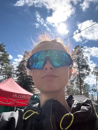 A selfie taken in a natural outdoor setting, possibly a park or wooded area, with the subject looking upward. The subject is wearing reflective goggles that obscure the eyes and reflect the surrounding trees and sky. Part of a red canopy or tent is visible in the lower portion of the frame, with the website "vk.com/fs" printed on it. The subject is wearing a dark-colored garment with yellow accents. The sky is blue with scattered white clouds and bright sunlight. The picture may be taken in Lesnoye, Russia, though there is no visible evidence to confirm the location.