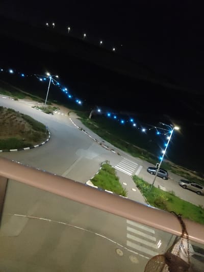 A nighttime perspective from a balcony overlooking a lit road. String lights with blue bulbs illuminate the road and nearby greenery. Street lights add further brightness. A crosswalk marks a pedestrian area. Two cars are parked near a grassy patch. The view from the balcony includes a glimpse of an object hanging off to the right of the balcony's railings.
