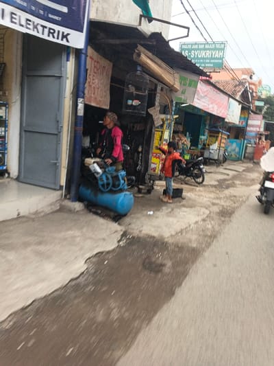 The image captures a street scene in Cangkuang Kulon, Indonesia. The foreground consists of an uneven concrete sidewalk on the left, transitioning to an asphalt road on the right, both exhibiting wet patches and scattered debris.

On the left, a building with a white banner displaying "ELEKTRIK" and a grey metal double door is partially visible. Immediately adjacent, an open-fronted commercial establishment features a sign reading "TERIMA SEJU MOTOR" (likely indicating motorcycle service) and a hanging birdcage. A blue air compressor rests on the sidewalk directly in front of this stall.

An adult individual, possibly female, wearing a pink jacket and a head covering, stands within the doorway of the open-fronted stall, looking towards the right. To her right, a young child, dressed in an orange and black jacket, light blue jeans, and brown shoes, stands on the sidewalk with both arms extended and pointing towards the right.

Further along the street, multiple other shops or stalls with various signs are present. A prominent green sign overhead reads "MASJID JAMI DAN MAJLIS TA'LIM AS-SYUKRIYAH." A black motorcycle is parked on the sidewalk near the child. Towards the right side of the frame, a partially visible motorcycle with a rider appears to be moving away from the viewer on the road. Overhead electrical wires are visible against an overcast sky.