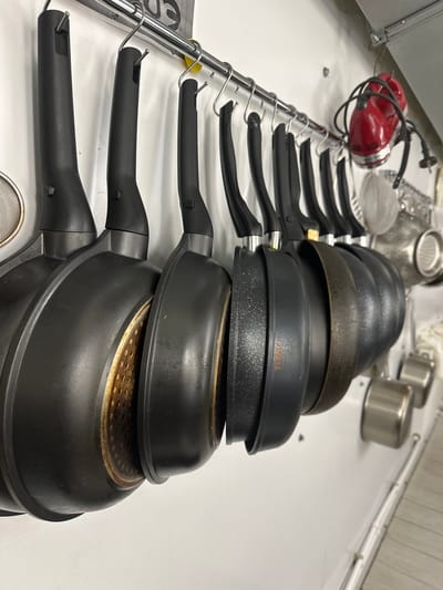 A row of black frying pans, stacked on top of each other, are positioned on a white surface. The pans have black handles and are hanging from a silver metal rack. A red hand mixer and various metal cooking utensils, including small pots and a strainer, are also present on the surface. The location is Новоглаголево, Russia.