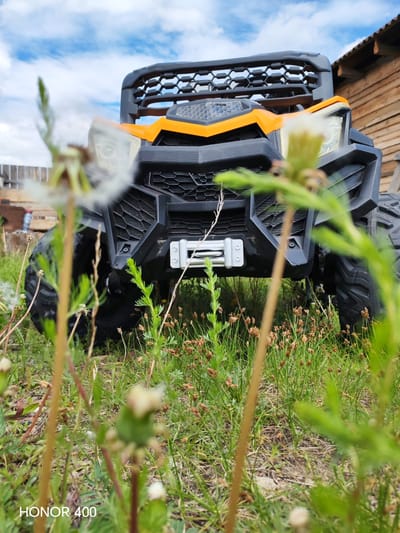 An eye-level, low-angle medium shot captures the front of a black and orange ATV parked outdoors. The ATV is the primary subject, positioned in the center of the frame and taking up a significant portion of the image. Its grille, headlights, and front bumper are clearly visible. A silver winch is mounted on the front of the ATV. In the foreground, out-of-focus green grass and plants with some dried seed heads create a natural, unmanicured environment. The background features a wooden building with a corrugated metal roof on the right side and a wooden fence on the left. The sky above is blue with scattered white clouds. The image is an outdoor scene in what appears to be a rural or semi-rural setting. The location is identified as a village in Russia.