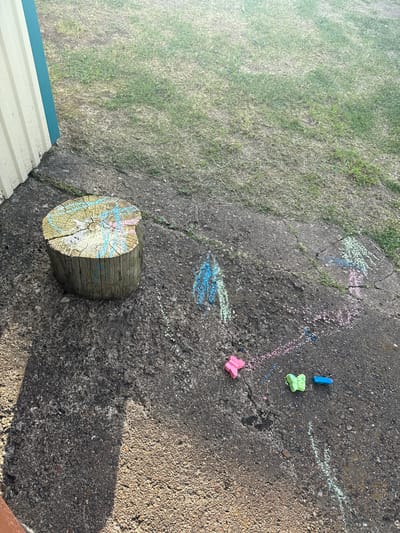 The image shows a wooden tree stump on a cracked concrete surface with chalk drawings and a few small plastic toys. The stump is decorated with colorful chalk markings, primarily in blue and pink. Chalk lines in various colors, including blue, pink, and green, are scattered on the concrete around the stump. Two plastic toys, one shaped like a pink butterfly and another like a green butterfly, are visible on the ground, along with a small blue rectangular object. In the background, dry grass and a portion of a building with yellow siding and a blue trim are visible. The lighting suggests it is daytime. The scene is located outdoors, on what appears to be a paved area adjacent to a grassy section.