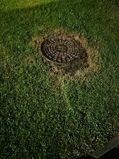 A manhole cover with a concentric circular pattern is visible in the center of a patch of brown, dead grass, surrounded by healthy green grass. The lighting suggests it is nighttime, possibly illuminated by a spotlight or streetlight, casting a shadow. The image was captured in Pabradė, Lithuania. There are no people or animals present in the scene. The environment is a grassy area, likely a park or roadside.