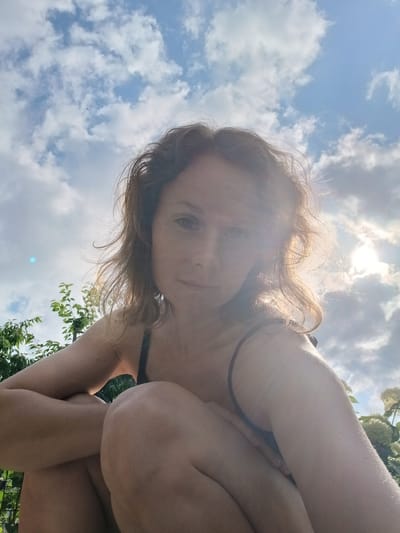 In Maslova Pristan', Russia, a woman with auburn hair is captured in a low-angle selfie outdoors. She is crouching, with her arms and legs pulled in towards her body. The woman is wearing a dark-colored top, possibly a bikini top. Her hair is windswept, and the sunlight is glinting off her face and hair, creating a lens flare effect. Behind her, the sky is filled with a mix of blue sky and white clouds. Lush green foliage, including trees and some white flowers, is visible to the left and right of the woman, suggesting she is in a garden or park setting. The overall atmosphere is bright and natural, with the sun being a prominent element in the background.