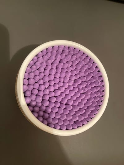 A top-down, medium shot captures a round, white container filled with many purple cotton swabs arranged in a dense, honeycomb-like pattern. The container is placed on a dark, matte surface, likely a table, which casts a soft shadow to the left of the container. The background is a plain, dark gray. The lighting is subdued, highlighting the texture of the cotton swabs and the smooth surface of the container. There are no people or other objects visible in the frame. The location is in the city of Милицейский посёлок, Russia.