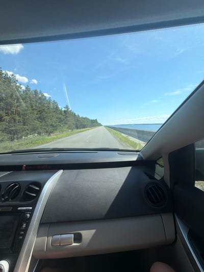 The image shows a view from the interior of a car driving along a road. The road is paved and has lane markings. On the left side of the road, there is a dense forest of pine trees. On the right side of the road, there is a large body of water, likely a reservoir or a river, with a concrete barrier separating the road from the water. The sky is blue with some scattered white clouds and a faint contrail from an aircraft. The dashboard of the car is visible in the foreground, with parts of the center console and air vents. The overall scene suggests a sunny day, possibly in the summer, with the car traveling along a scenic route. While the image is taken from a car, there is no specific information within the image to confirm the location as Vyshhorod, Ukraine, or any other specific city.