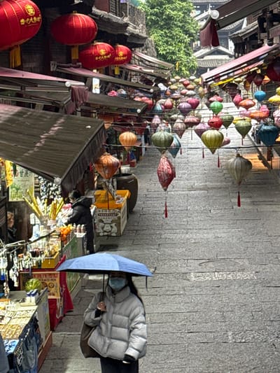 The image depicts an uphill, stone-paved street within a market area in Pengshui Miao and Tujia Autonomous County, China. Numerous lanterns are strung overhead; these include spherical red lanterns with yellow tassels displaying Chinese characters and a canopy of multicolored, teardrop-shaped fabric lanterns with red tassels. Traditional Chinese architectural elements, including dark-tiled roofs and wooden structures, line both sides of the street, with dark awnings extending over various shop fronts.
In the mid-foreground, a female subject is walking uphill, holding a light blue umbrella. She wears a light-colored puffer jacket, dark trousers, dark gloves, and a light blue surgical mask. A dark shoulder bag is carried on her right side. To her left, at a market stall, another individual in a dark puffer jacket is visible, appearing to tend to goods that include fresh fruit and items on sticks. Further up the street, additional indistinct figures are present amidst various stalls displaying merchandise and signage with Chinese characters, one prominent sign reading "甘蔗汁" (sugarcane juice). The street surface is composed of grey, rectangular stone blocks. Green foliage is visible in the upper background, indicating a natural element beyond the immediate structures.