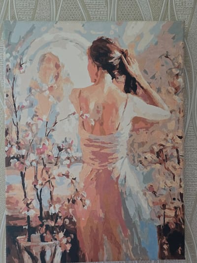 The image shows a painting of a person lying down, possibly on a bed or in a field of flowers, with a white cloth draped over them. The painting uses soft, pastel colors, with shades of pink, peach, and light blue dominating the scene. The person's hair is dark, and their face is partially obscured. There are also elements that suggest a natural setting, such as branches with small pink and white flowers, resembling cherry blossoms. The overall impression is one of tranquility and gentle beauty. The painting is rendered in a style that appears to be paint-by-numbers, with distinct sections of color that suggest the artist followed a pre-drawn outline. The painting is set against a textured background, possibly a wallpaper or fabric. There is no visible text in the image.