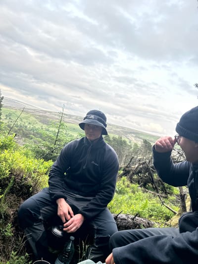 Two young men, both wearing black clothing, are sitting on a log in a forest clearing. The man on the left is wearing a black bucket hat and is holding a black metal cup and a clear water bottle. The man on the right is wearing a black beanie and is holding his hand to his face. Both men are looking in different directions. The clearing is overgrown with green plants and some branches and logs. There is a distant view of a valley with green hills in the background. The sky is cloudy. This scene is in Macclesfield Forest, UK, at coordinates 53.26 latitude and -1.99 longitude.