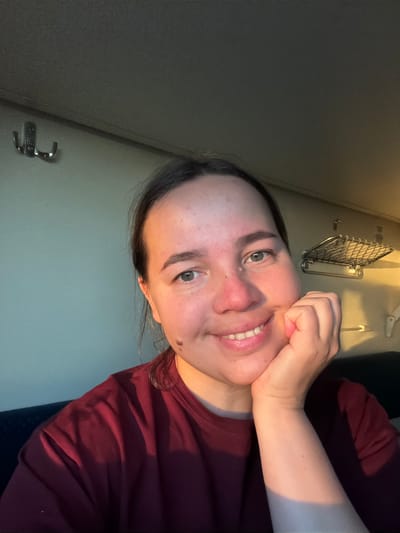 A woman is posing for a selfie, tilting her head to the side and smiling. She is wearing a burgundy top. Her hand is touching her face. Behind her is an interior setting, possibly a train compartment or a small room. There is a luggage rack and some fixtures on the wall. The scene appears to be illuminated by natural light coming from the side. The location in the city of Kizner, Russia, cannot be verified based solely on this image.