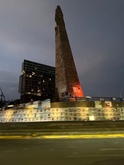 The image depicts an urban night scene in Guadalajara, Mexico, focusing on the iconic "Los Arcos" monument. The stone arch of Los Arcos stretches diagonally across the frame, with various posters and flyers affixed to its surface. Adjacent to the monument is a modern high-rise building with illuminated windows, suggesting residential or commercial occupancy. A street runs along the side, its surface reflecting ambient light, and a wall covered in posters stands near the road. The sky is a dark, muted blue, indicating nighttime. Construction equipment, specifically a crane, is visible in the lower right corner, suggesting nearby development activities. There are no people visible in the photograph.