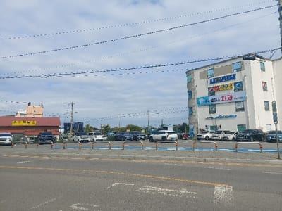 This image depicts a parking lot and two commercial buildings under a partly cloudy sky in 강화군, South Korea. In the foreground, an asphalt road with faded white and yellow lane markings is visible, bordered by a row of blue and silver bollards. Beyond the bollards, a dirt and gravel parking area contains approximately ten parked vehicles, including several white SUVs/cars, dark-colored SUVs, and a blue sedan/SUV.
To the left of the parking lot is a single-story or low-rise building with a reddish-brown roof, featuring a prominent yellow sign displaying Korean text "온수 제빵소" (Onsu Bakery). Further left, partially obscured, is a taller, light-colored building.
On the right side of the image, a multi-story, light-colored commercial building with a rounded corner dominates the scene. Its facade is adorned with multiple large signs, primarily in blue and white, displaying Korean text and graphics. A prominent sign on the second floor clearly indicates "치과" (Dental Clinic). Utility poles and power lines crisscross the upper portion of the sky. No individuals are clearly discernible in the scene.
