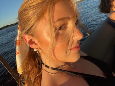 A close-up shot features a light-skinned woman with reddish-blonde hair on a boat on a river. She has a thin black choker around her neck and another gold necklace below. She is wearing pearl earrings and a scarf tied to her hair. The river water is visible in the background with trees on the bank in the distance. The location appears to be the Zambezi River near Victoria Falls, Zimbabwe. Another person is partially visible seated beside the woman.