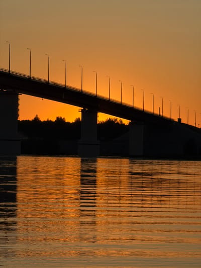 The image shows a bridge over a river at sunset, casting reflections on the water. The sky is a gradient of orange and yellow, with a silhouette of trees on the horizon. The bridge is lined with streetlights, which are also silhouetted against the sky. The water is calm, with gentle ripples reflecting the colors of the sunset and the bridge. The image captures a serene and picturesque moment.