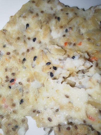 A close-up, high-magnification view reveals a prepared food item positioned on an indistinct light-colored surface in Oba-Aboji, Nigeria. The primary subject is an irregularly shaped mass composed of an opaque, off-white to pale yellowish matrix. Embedded extensively within this matrix are numerous small, dark brown to black ovoid seeds. Also visible are lighter-colored, irregular granular fragments, possibly oat flakes or cereal grains, along with occasional small, vibrant reddish-orange particulate matter. Scattered within the food item are several thin, translucent, hair-like strands. The object's edges are uneven and slightly disrupted, with a distinct crevice or tear visible on the right side. No people, actions, or direct interactions are depicted in the frame.