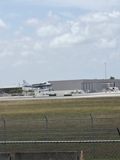 The image shows a seaplane in mid-air over a runway at an airport in Fort Lauderdale, United States. The seaplane, a white and blue Cessna, has the registration number N4215Y on its tail. It is being observed from behind a chain-link fence. In the background, there are several hangars and other aircraft on the tarmac, including two small white jets. The sky is partly cloudy. The scene appears to be a clear day at a general aviation airport.