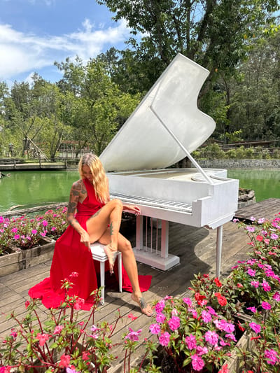 A blonde woman with tattoos is sitting on a stool in front of a white grand piano. She is wearing a long, flowing red dress that is split on the side, revealing her legs. The piano is positioned on a wooden deck next to a green pond. There are many pink and red flowers planted in wooden boxes around the deck. In the background, there are trees and a blue sky with white clouds. The location is Cam Ly - Da Lat, Vietnam.