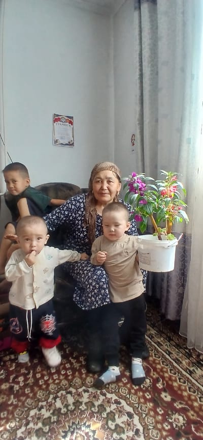 A middle-aged woman wearing a headscarf and a floral dress sits on a couch in a room. Two toddlers are in front of her. One toddler, wearing a light-colored shirt with animal prints and dark pants, stands to the left, with their hand to their mouth. The other toddler, in a tan shirt and black pants, stands to the right, holding a white bucket with a plant in it. Behind the woman, a young boy in a green shirt sits on the couch. A certificate is hung on the wall behind the woman. The room has light-colored walls, a patterned rug on the floor, and sheer curtains. The scene appears to be an indoor family gathering.