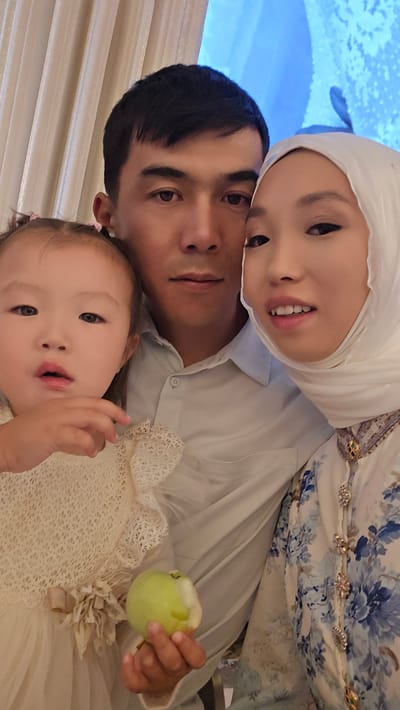 The image shows a family selfie, with three people close together. In the foreground, a young woman wearing a white hijab and a patterned dress with blue floral designs is looking at the camera, smiling. Next to her, a man in a light-colored shirt is also looking towards the camera, with a neutral expression. Behind them, a baby with light skin and dark eyes is nestled between the man and the woman, also looking towards the camera. The baby is wearing a cream-colored lace dress and holding a partially eaten green apple in its left hand. The background appears to be indoors, with light-colored curtains or blinds visible. The lighting suggests it could be daytime, though the exact time of day is not discernible. The overall mood of the photo seems warm and familial. No visible text is present in the image. The location context provided is Karakol City, Kyrgyzstan.