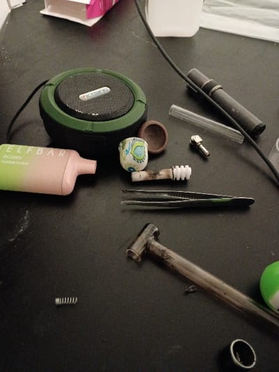 The image shows a variety of objects scattered on a dark grey surface, likely a table, in a residential setting.  The objects include a green portable speaker, a pink and green disposable vape pen, a metal spring, a pair of tweezers, a small brown ceramic bowl, a glass tube, and a small metal object with a black ring. The location is an apartment or home in Seattle, Washington, United States, based on the provided coordinates. The scene appears to be a still life, with no people or animals present.