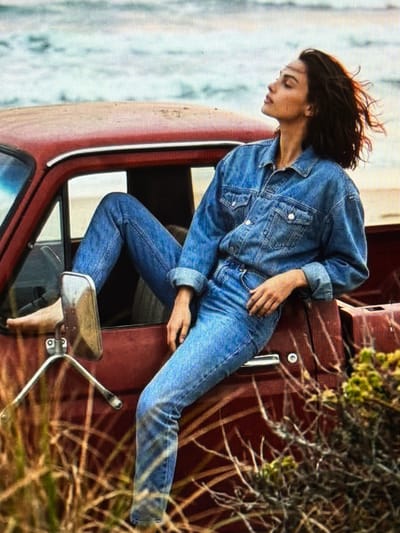 A woman with dark hair is sitting on the edge of the bed of a rusty red pickup truck. She is wearing a denim jacket and jeans, and her head is tilted back with her eyes closed, as if enjoying the sun. Her legs are bent, with one foot resting on the side mirror. The truck is parked on a sandy area with dry grass and bushes in the foreground. In the background, a body of water, likely the sea or a large lake, is visible under a cloudy sky. The scene appears to be a quiet, natural setting, possibly a beach or a remote coastal area. There is no indication of Bobrovo, Russia in the image.
